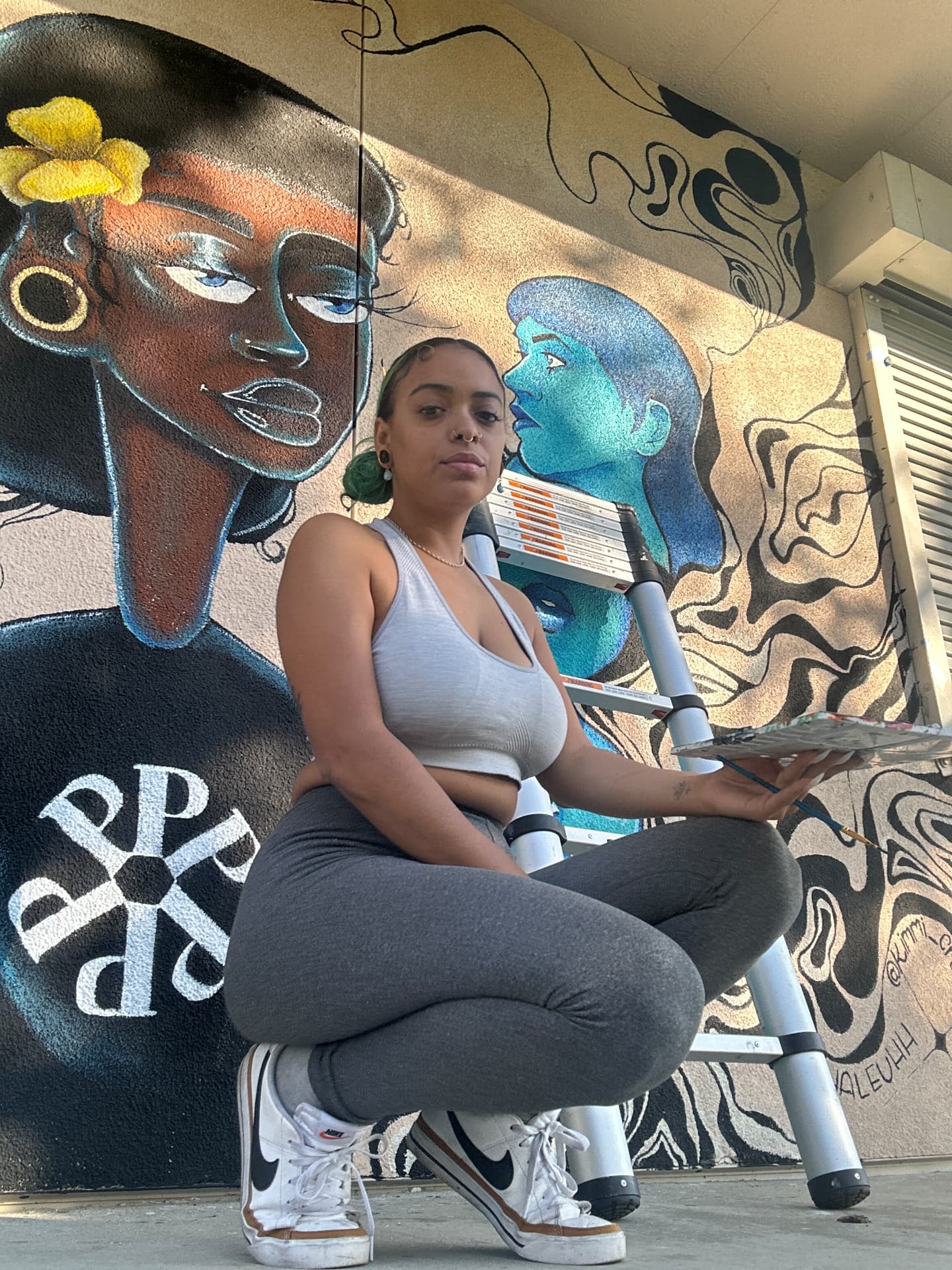 Chaleuh crouching with her palette and brush in front of a mural of blue faces and flowing lines